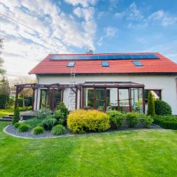 Dom jednorodzinny z czerwonym dachem, panelami słonecznymi, ogrodem i tarasem, drabina oparta o dach.