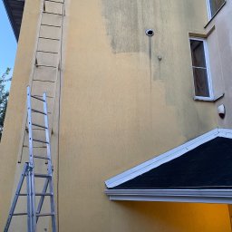 Jaksiepatrzy24 - Żółta elewacja budynku z widocznymi śladami zabrudzeń i częściowo wyczyszczoną powierzchnią, oparta o ścianę aluminiowa drabina rozstawna, fragment dachu i okna.