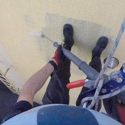 Jaksiepatrzy24 - Pracownik w uprzęży alpinistycznej, z perspektywy pierwszej osoby, czyści elewację budynku za pomocą myjki ciśnieniowej, widoczne fragmenty umytej i brudnej powierzchni.