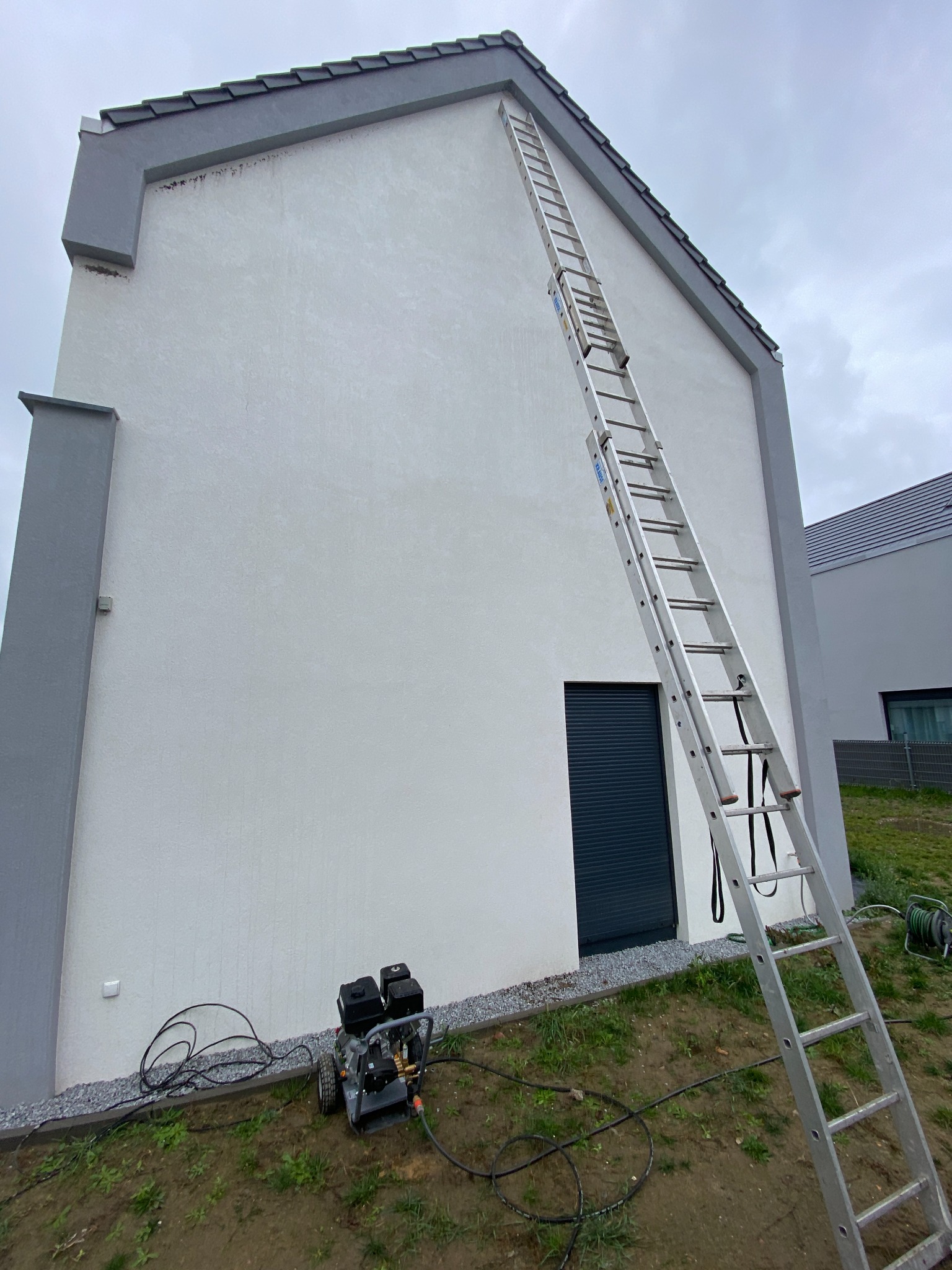 Aluminiowa drabina oparta o ścianę budynku z jasną elewacją, widoczna myjka ciśnieniowa i ciemne rolety okienne.