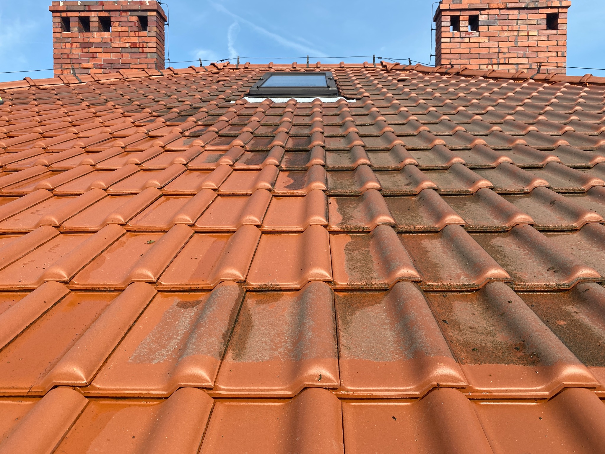 Częściowo umyta, czerwona dachówka z widocznymi smugami brudu, okno dachowe i dwa ceglane kominy na tle błękitnego nieba.