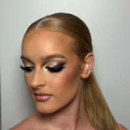 Portret kobiety z intensywnym, wieczorowym wizażem: złote cienie, czarny eyeliner i sztuczne rzęsy. Włosy gładko zaczesane do tyłu w niski kucyk, widoczne kolczyki w uchu.