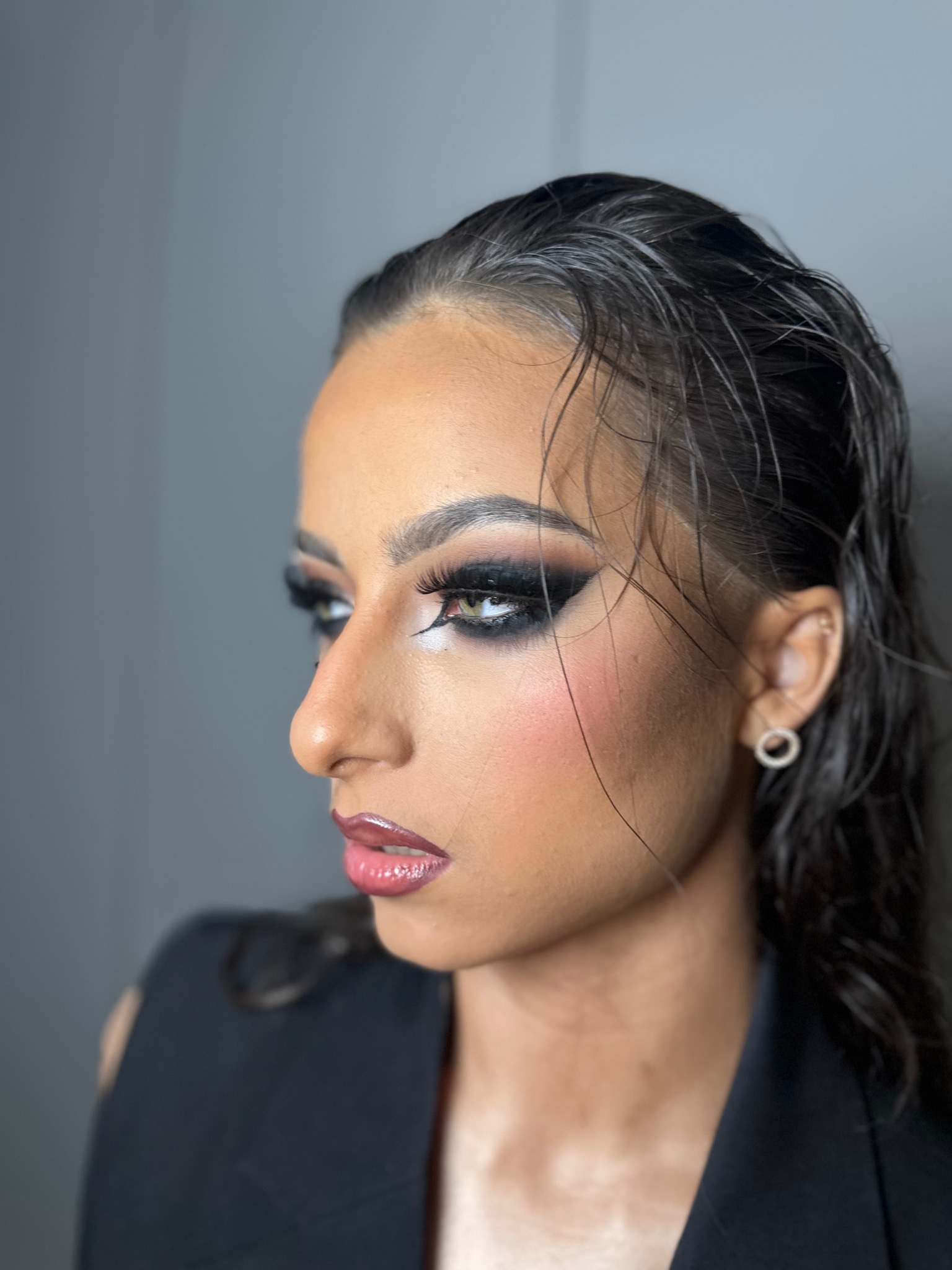 Profil kobiety z intensywnym makijażem smoky eye, błyszczącymi ustami i mokrymi włosami, prezentującym efektowny look na szaro-niebieskim tle.