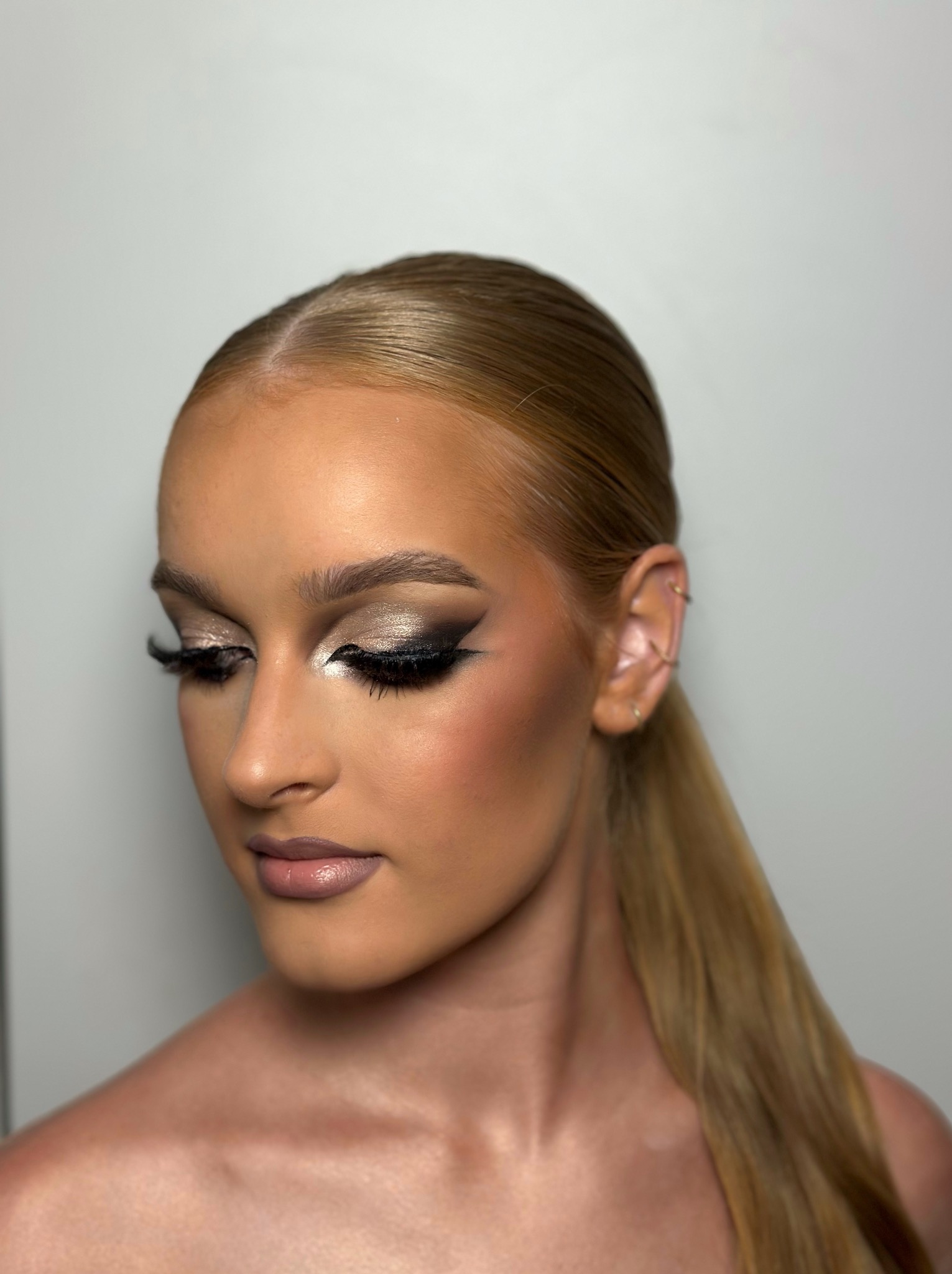 Portret kobiety z intensywnym, wieczorowym wizażem: złote cienie, czarny eyeliner i sztuczne rzęsy. Włosy gładko zaczesane do tyłu w niski kucyk, widoczne kolczyki w uchu.