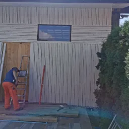 Pracownik w pomarańczowych spodniach montuje pionowe deski na ścianie budynku, używając wkrętarki akumulatorowej; widoczna drabina, poziomica i częściowo ułożona kostka brukowa wokół basenu.