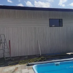 Ściana domu w trakcie budowy z drewnianą elewacją, widoczne rusztowania, basen i niebieskie niebo.