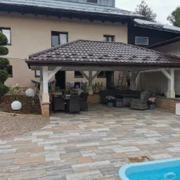 Zadaszony taras z meblami ogrodowymi i basenem, otoczony kostką brukową w różnych odcieniach szarości i beżu, z widocznym fragmentem żółtego węża ogrodowego i ozdobnym drzewkiem.