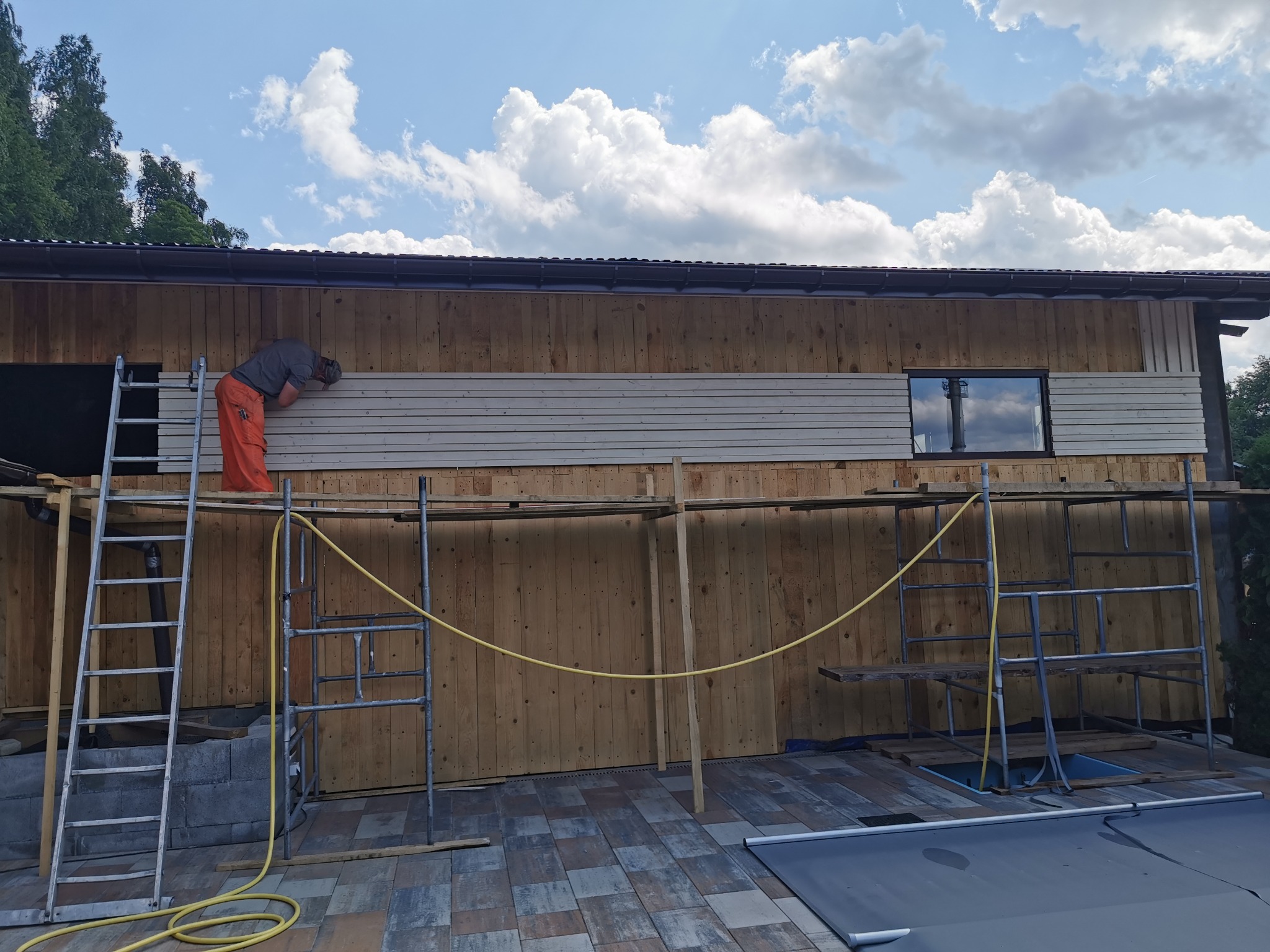 Pracownik w pomarańczowych spodniach montuje jasne panele na drewnianej elewacji budynku, widoczne rusztowania i drabina, błękitne niebo w tle.