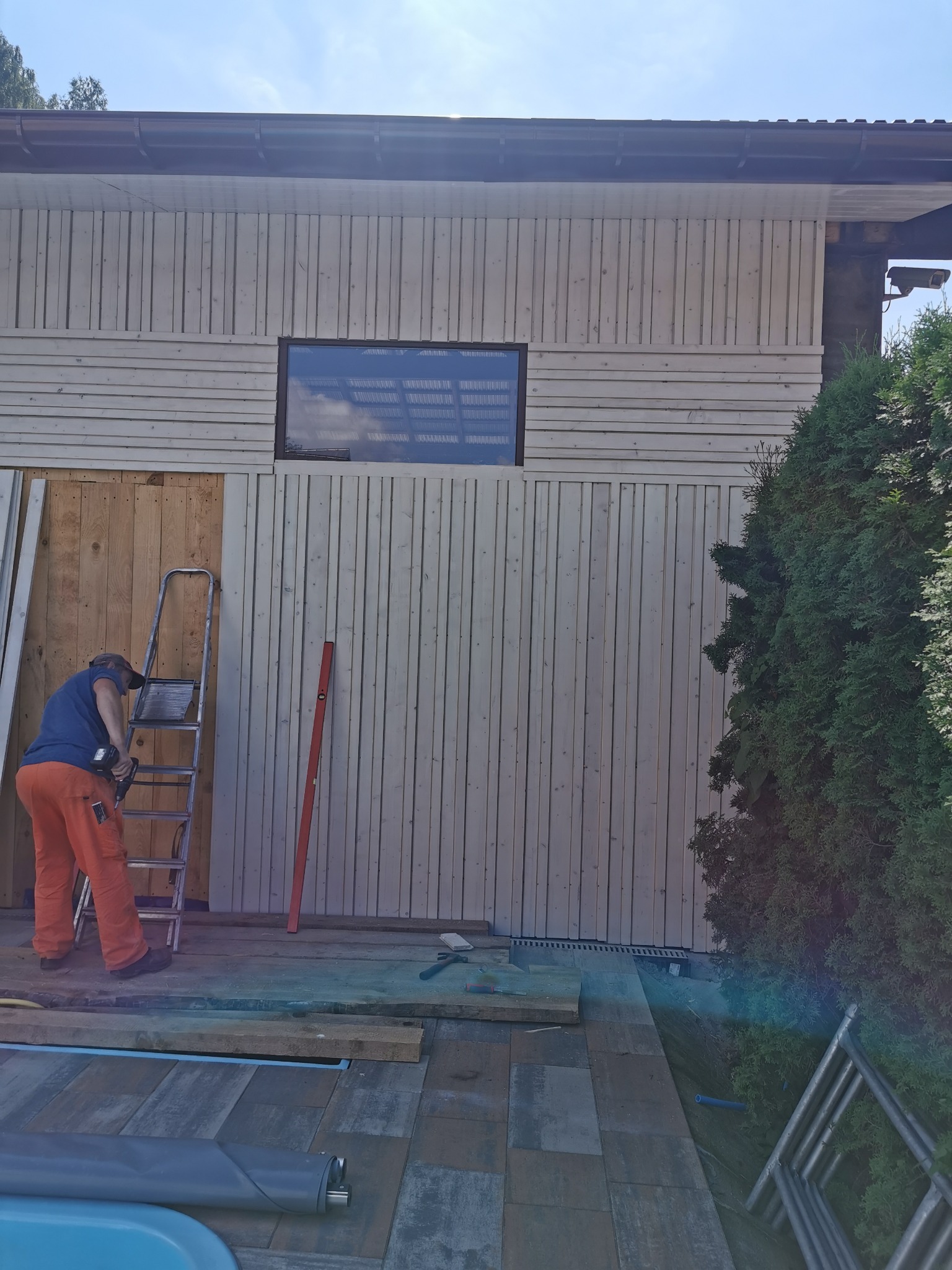 Pracownik w pomarańczowych spodniach montuje pionowe deski na ścianie budynku, używając wkrętarki akumulatorowej; widoczna drabina, poziomica i częściowo ułożona kostka brukowa wokół basenu.