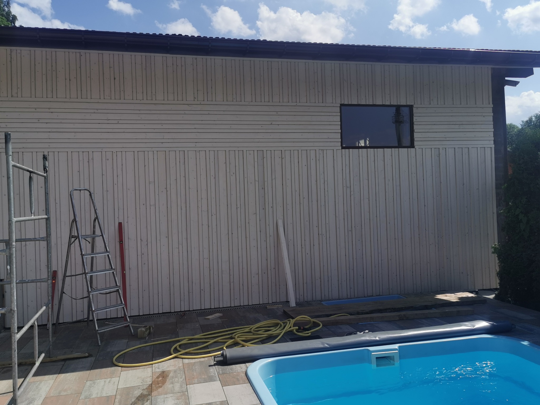 Ściana domu w trakcie budowy z drewnianą elewacją, widoczne rusztowania, basen i niebieskie niebo.