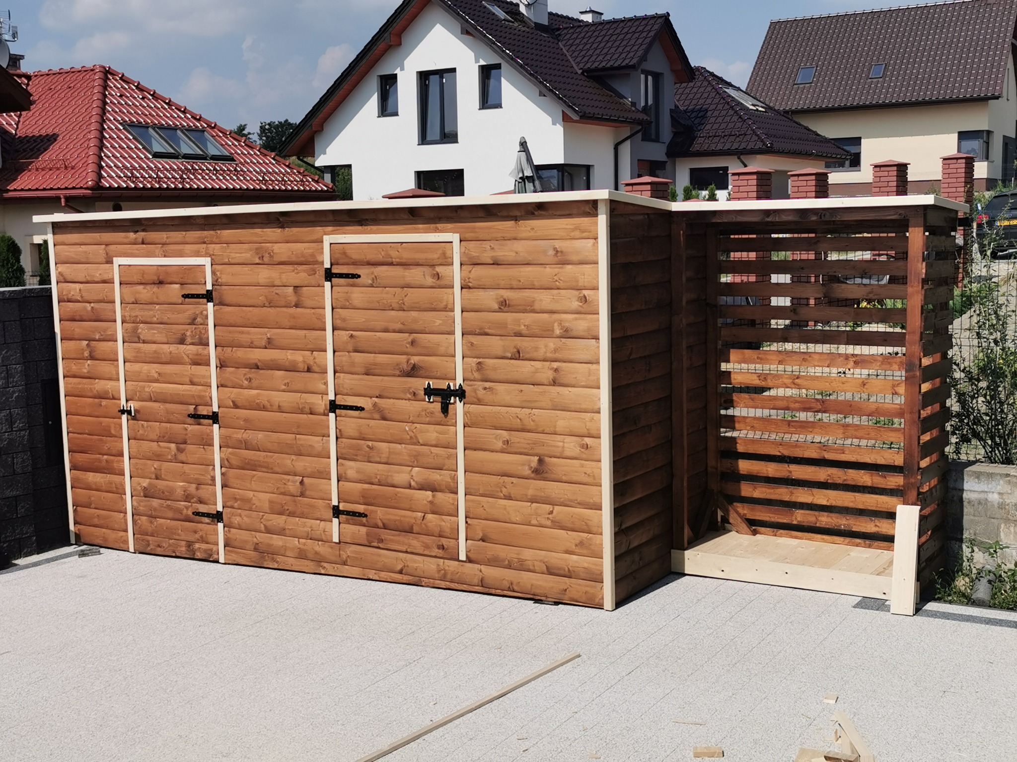 Drewniana wiata z trzema drzwiami i ażurową częścią na śmietnik, na tle domów jednorodzinnych z różnymi pokryciami dachowymi.