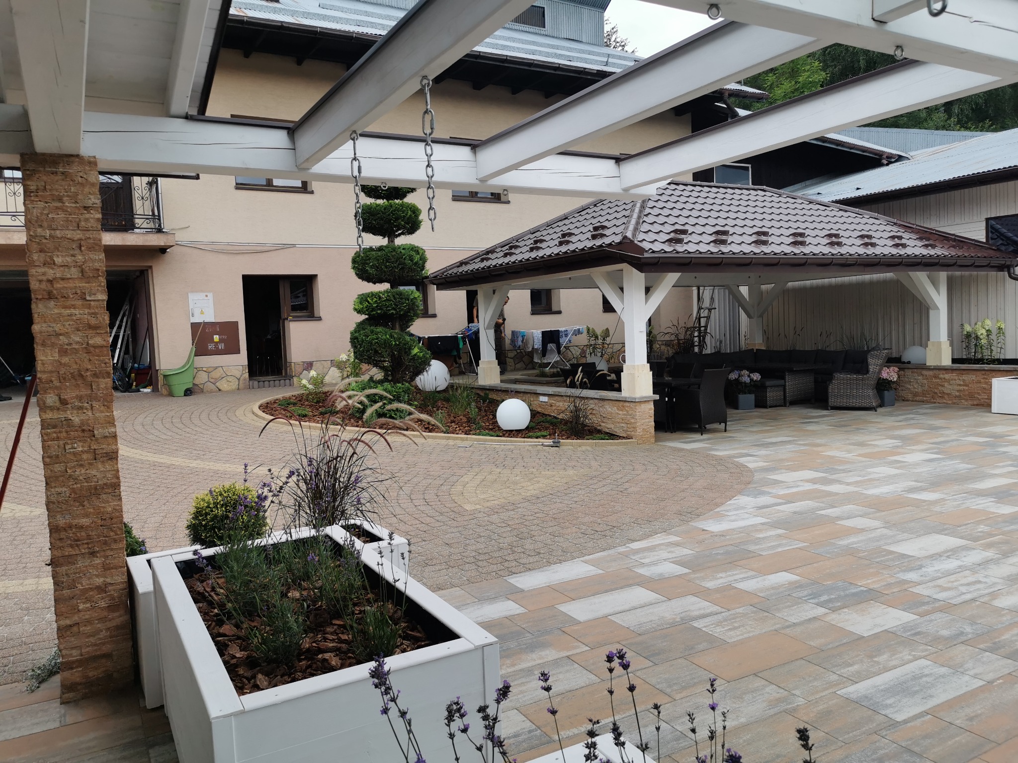 Przestronny dziedziniec z kostki brukowej w odcieniach beżu i szarości, z dekoracyjnymi roślinami w białych donicach, oświetlony kulami i z zadaszoną altaną wypoczynkową w tle.