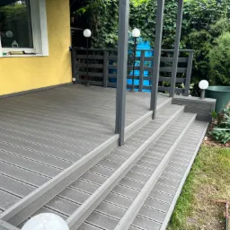 Szary taras kompozytowy z trzema schodami w Strzeniówce, otoczony zielenią, z widoczną lampą kulistą i fragmentem elewacji budynku w kolorze żółtym.