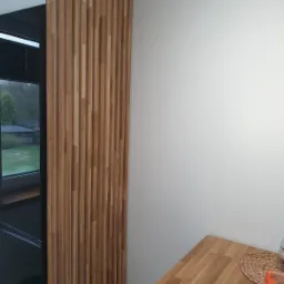 Drewniana ścianka działowa z pionowych listew w jasnym odcieniu drewna, oddzielająca kuchnię od salonu, widoczna z perspektywy blatu kuchennego.