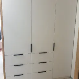 Biała szafa wnękowa z czarnymi, minimalistycznymi uchwytami, wykonana na wymiar w Strzeni&oacute;wce, z widocznymi sześcioma szufladami i trzema drzwiami.