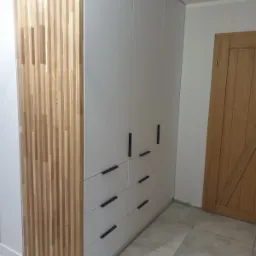 Biała szafa na wymiar z szufladami i drewnianymi lamelami po lewej stronie, czarne uchwyty, jasna podłoga, drzwi w tle.