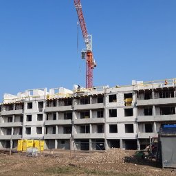 Grain - Wznoszenie surowego stanu wielopiętrowego budynku mieszkalnego z betonu komórkowego, widoczny żuraw budowlany i materiały budowlane na placu budowy.