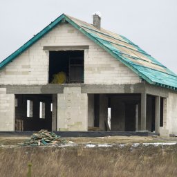 Grain - Surowy stan domu jednorodzinnego w trakcie budowy: ściany z bloczków Ytong, częściowo deskowane podłoże dachu z zieloną membraną, stos desek i paczka bloczków Ytong na działce.