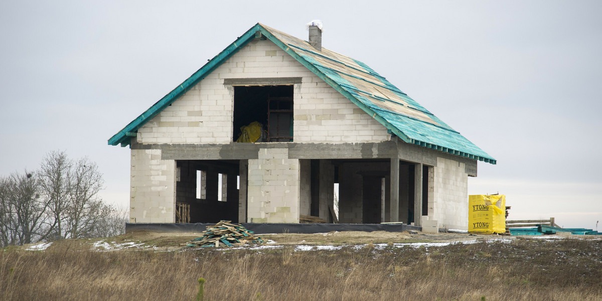 Surowy stan domu jednorodzinnego w trakcie budowy: ściany z bloczków Ytong, częściowo deskowane podłoże dachu z zieloną membraną, stos desek i paczka bloczków Ytong na działce.