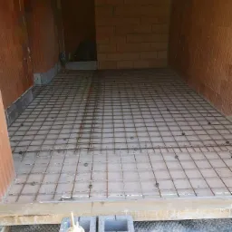 Przygotowanie zbrojenia pod wylewkę betonową wewnątrz budynku z cegły ceramicznej, widoczne bloczki betonowe i poziomica w dolnej części kadru.