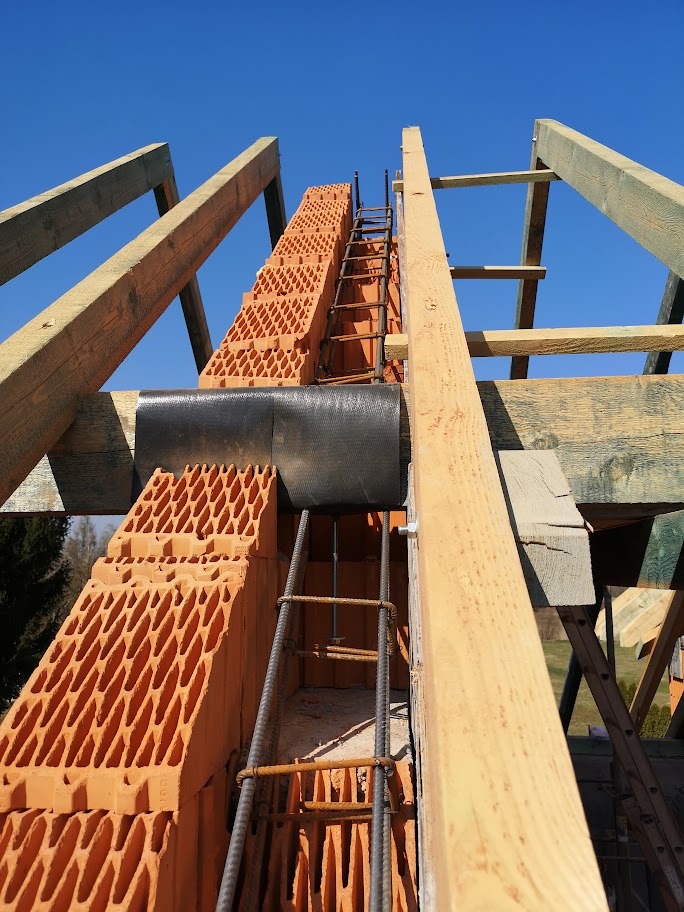 Wznoszenie ściany z pustaków ceramicznych Porotherm zbrojonej pionowo stalowymi prętami, widok od dołu na tle drewnianej konstrukcji dachu i błękitnego nieba.