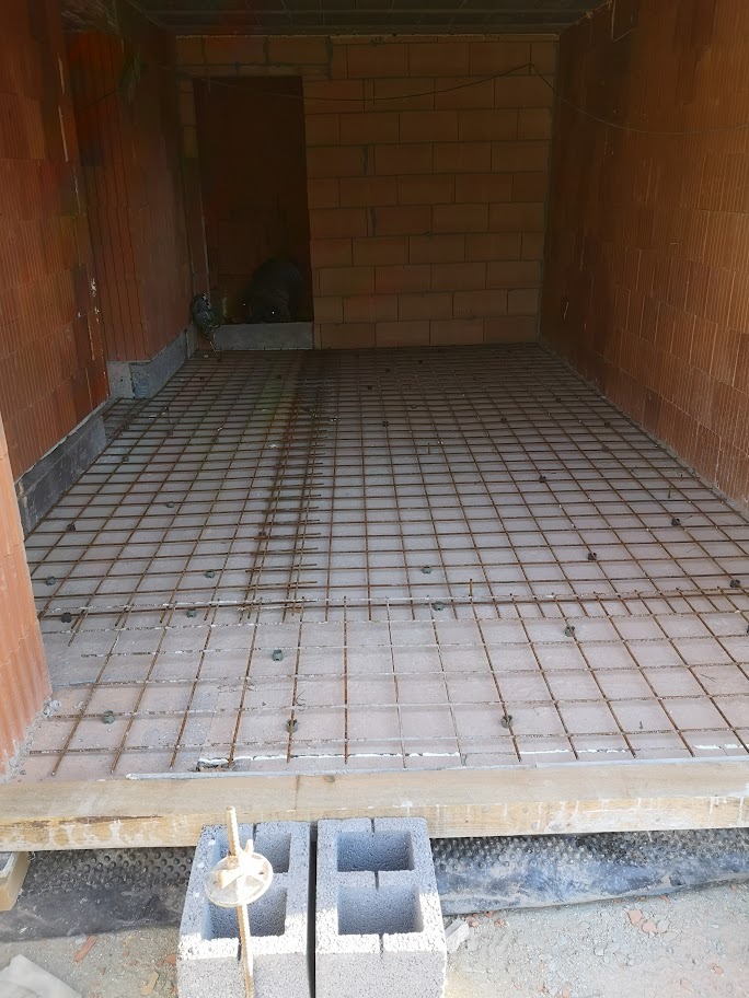 Przygotowanie zbrojenia pod wylewkę betonową wewnątrz budynku z cegły ceramicznej, widoczne bloczki betonowe i poziomica w dolnej części kadru.