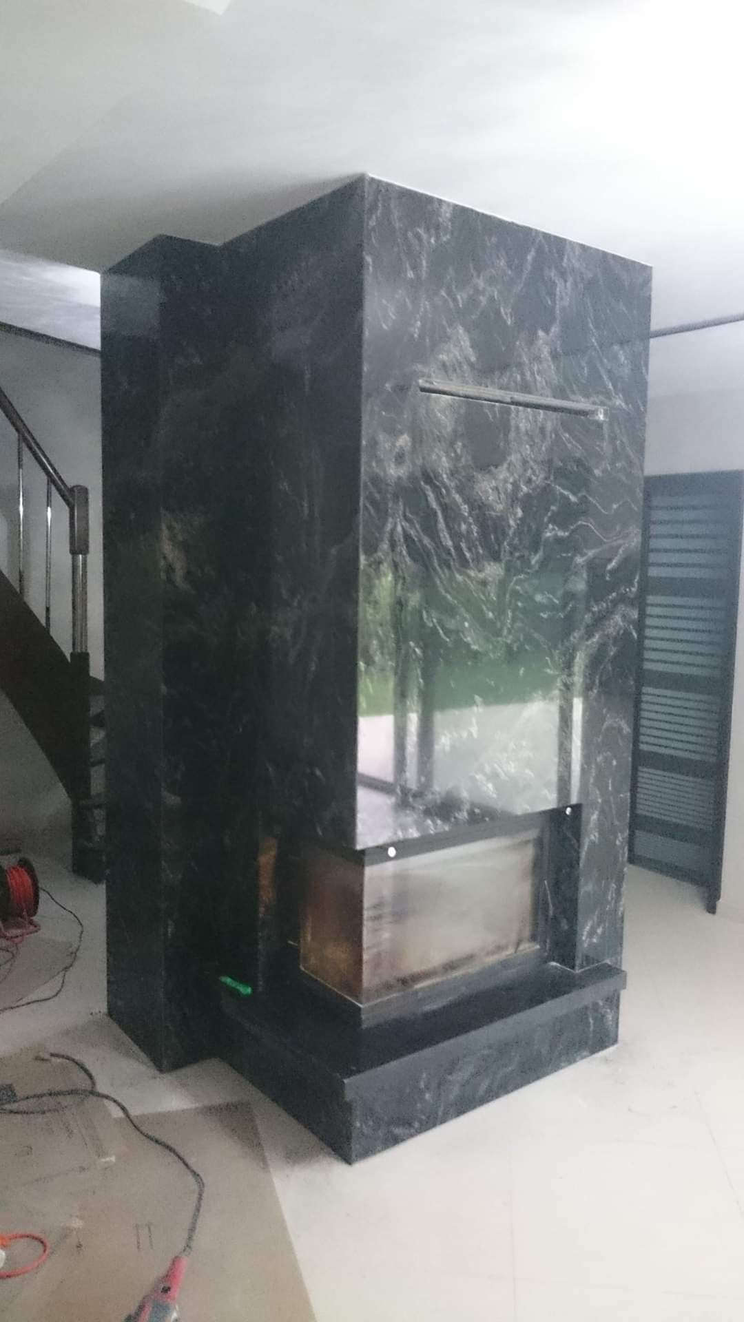 Obudowa kominka z czarnego kamienia z wyraźnym wzorem żyłkowania, widoczne schody w tle i przewody elektryczne na podłodze.