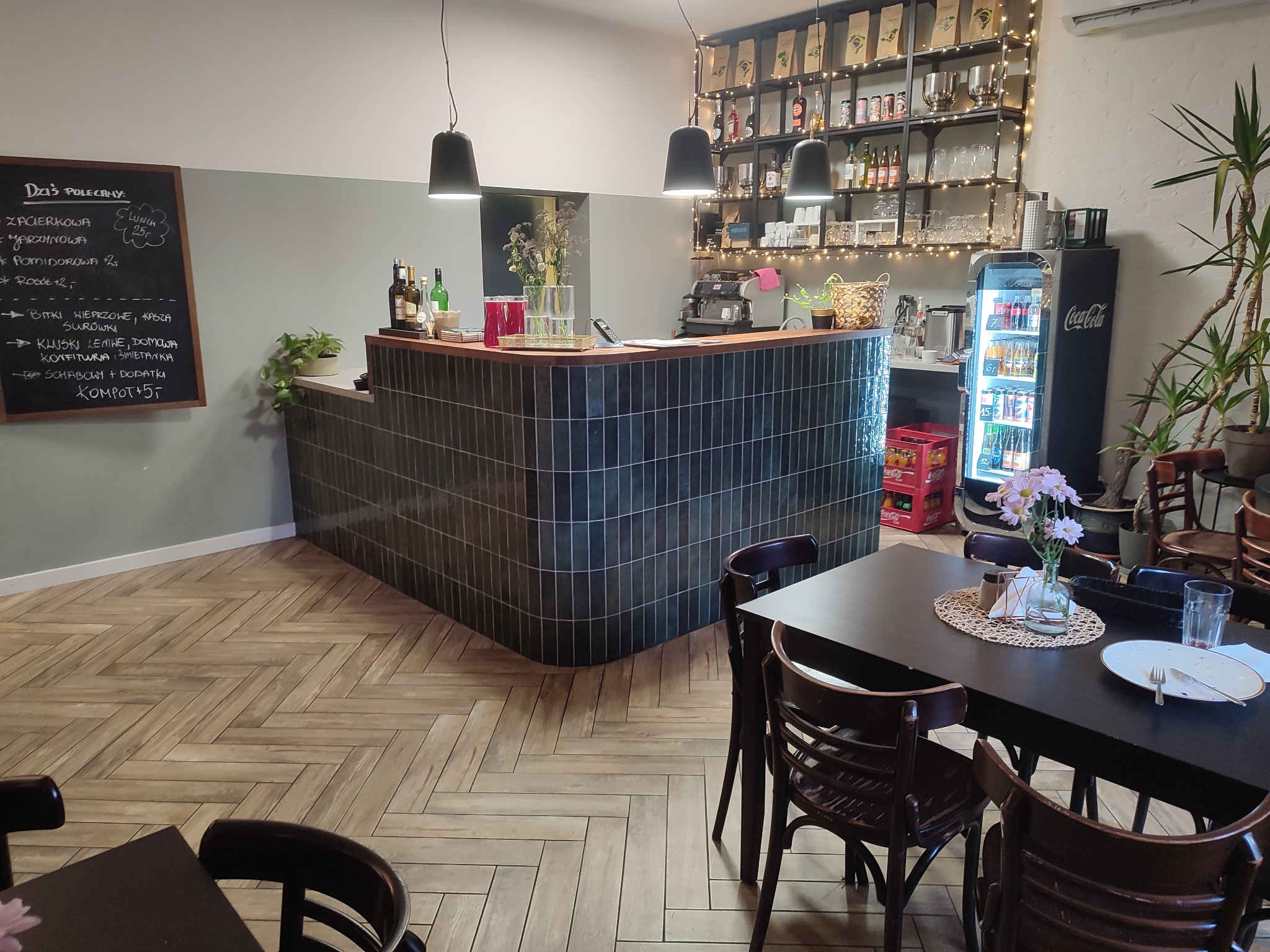 Wnętrze kawiarni z drewnianą podłogą ułożoną w jodełkę, bar z ciemnozielonymi płytkami, stoliki z ciemnymi krzesłami oraz regał z napojami i dekoracjami świetlnymi.