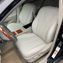 Jasnoszare skórzane fotele w samochodzie z drewnianą kierownicą i beżowymi dywanikami, widoczne detale wnętrza i naklejki informacyjne na drzwiach.