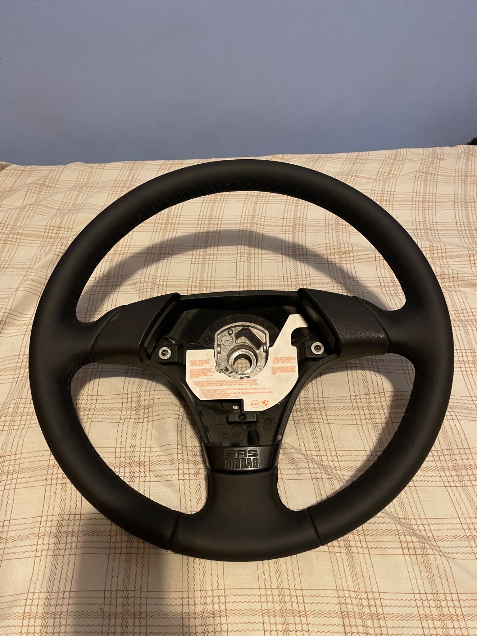 Czarna, skórzana kierownica samochodowa z widocznymi szwami, leżąca na wzorzystej narzucie, bez poduszki powietrznej.