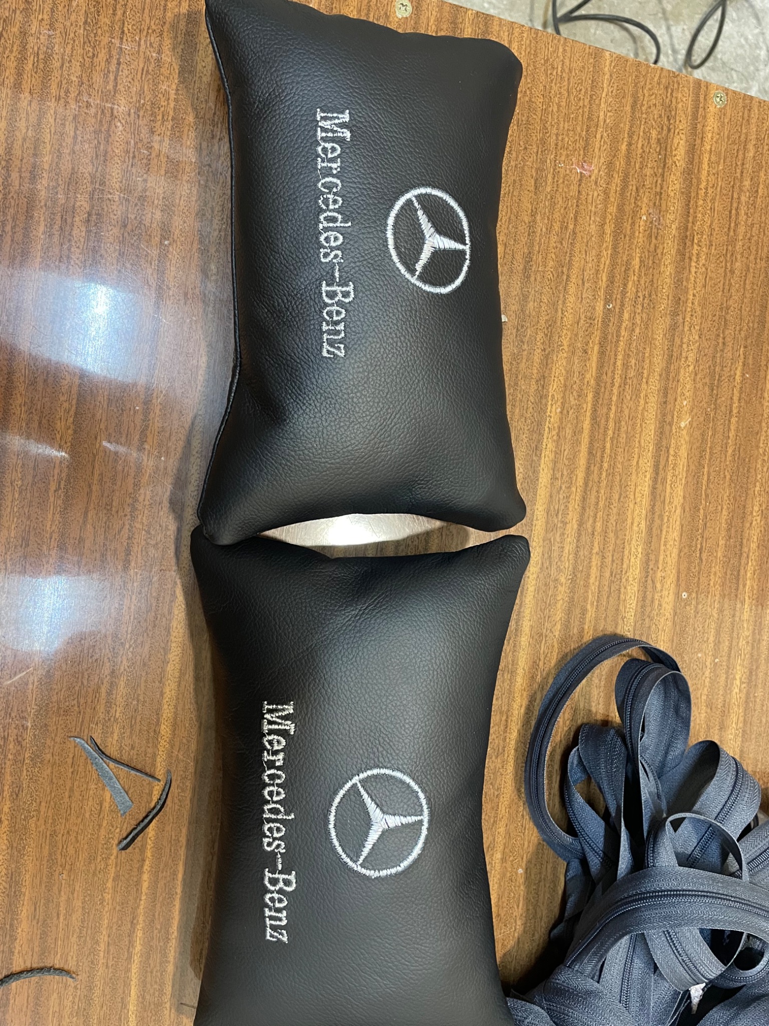 Dwie czarne, skórzane poduszki samochodowe z białym haftem logo Mercedes-Benz leżą na drewnianym stole, obok leżą resztki skórzanych ścinek i zwój czarnego zamka błyskawicznego.