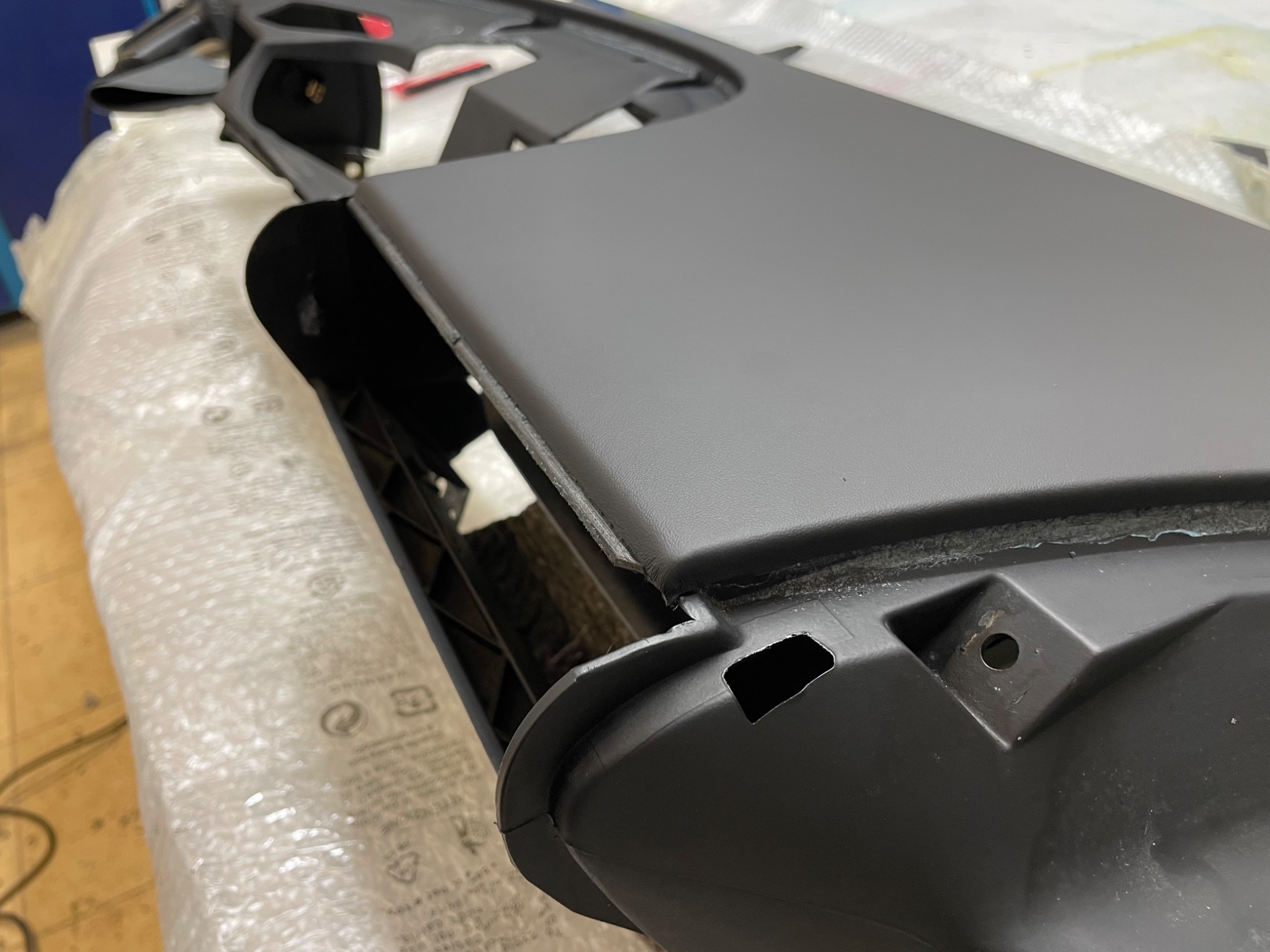 Szary, plastikowy element deski rozdzielczej samochodu, owinięty folią bąbelkową, widoczne otwory montażowe i krawędzie.