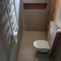 ProArt4you - Wykończona łazienka z beżowymi płytkami, wisząca toaleta, grzejnik drabinkowy z papierem toaletowym, wnęka z drewnianym wykończeniem i narzędzia na obudowie spłuczki.