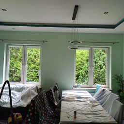 ProArt4you - Pokój przed odświeżeniem: ściany w odcieniach zieleni, meble zabezpieczone folią malarską, widoczny odkurzacz i puszka z farbą, okna z widokiem na zieleń.