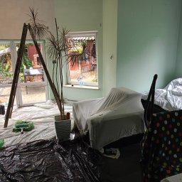 ProArt4you - Pokój w trakcie odświeżania: drabina oparta o okno, zabezpieczone meble pod białym materiałem, wiadro z farbą i wałek malarski na folii ochronnej, widoczna pomalowana na zielono ściana.