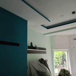 ProArt4you - Wnętrze w trakcie prac wykończeniowych: ściana pomalowana na intensywny morski kolor, sufit z dekoracyjnymi wnękami w tym samym odcieniu, zabezpieczone meble folią malarską, widoczna półka...