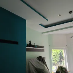 Wnętrze w trakcie prac wykończeniowych: ściana pomalowana na intensywny morski kolor, sufit z dekoracyjnymi wnękami w tym samym odcieniu, zabezpieczone meble folią malarską, widoczna półka...