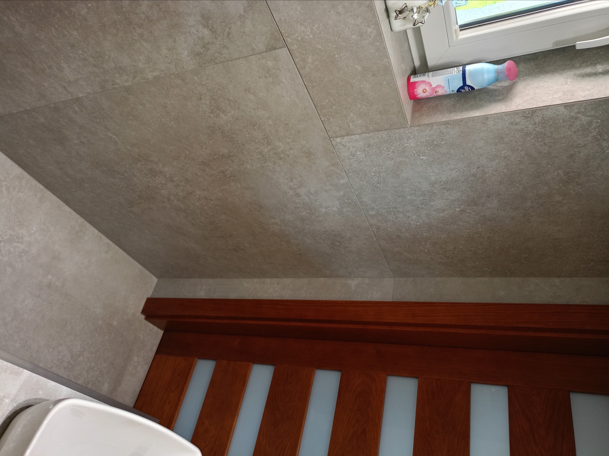 Wykończona ściana łazienki z szarych płytek, widoczna butelka odświeżacza powietrza na parapecie i fragment drewnianych drzwi z szybami.