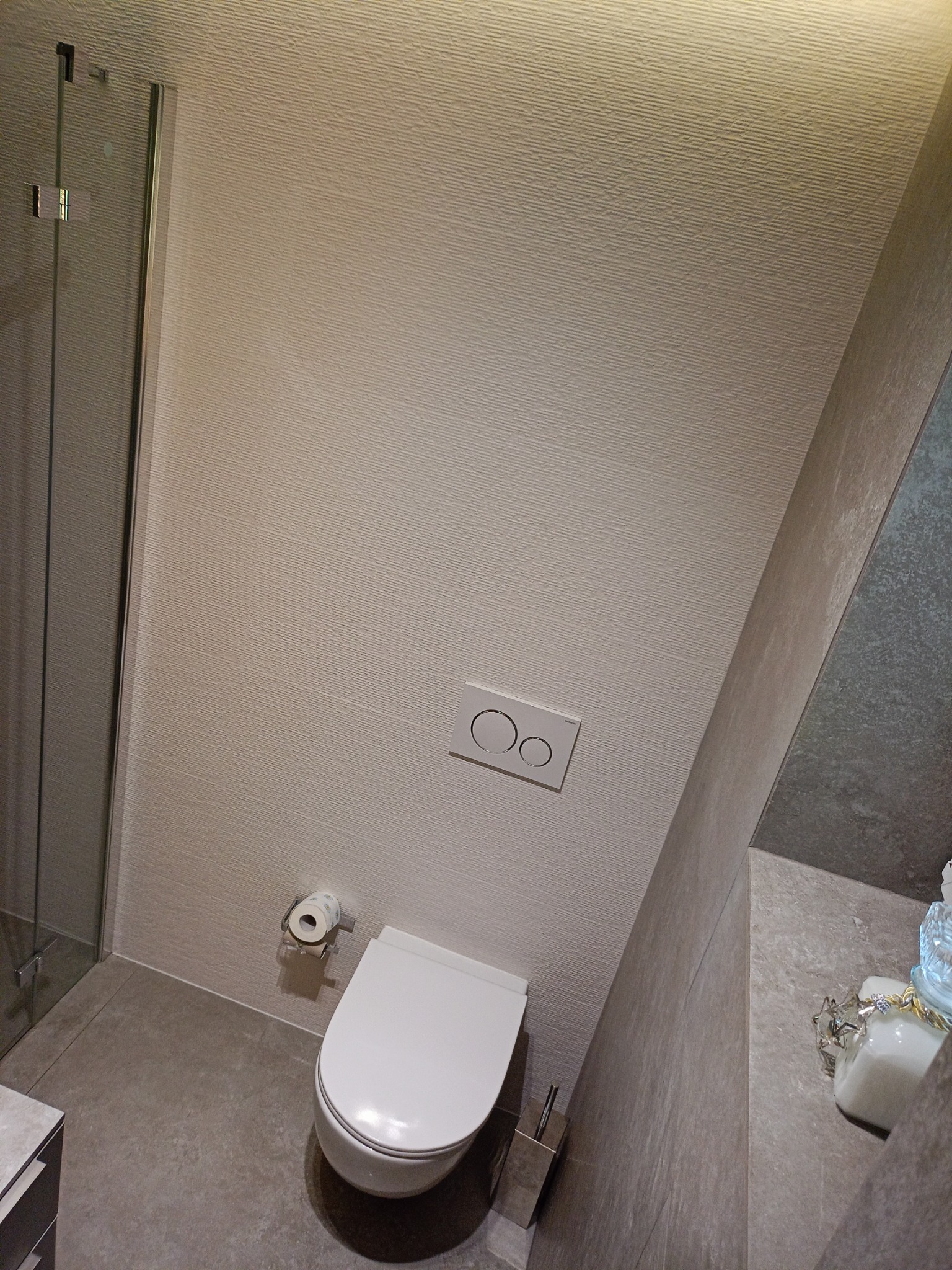 Nowoczesna łazienka z podwieszaną toaletą, szklaną kabiną prysznicową i teksturowaną ścianą w odcieniach beżu i szarości, uzupełniona o srebrny uchwyt na papier toaletowy i szczotkę do WC