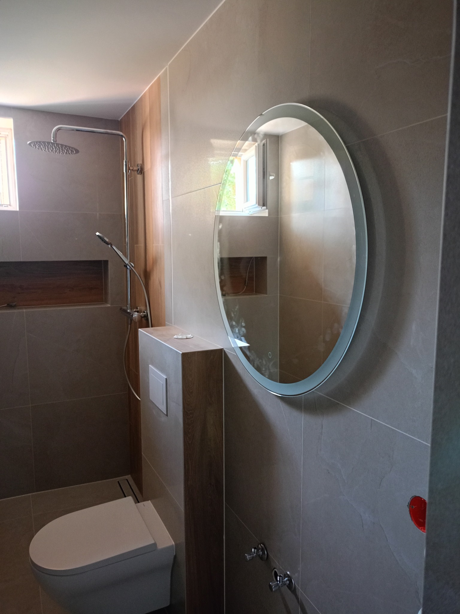 Nowoczesna łazienka w Częstochowie z szarymi płytkami, okrągłym lustrem z podświetleniem LED, podwieszaną toaletą i deszczownicą.