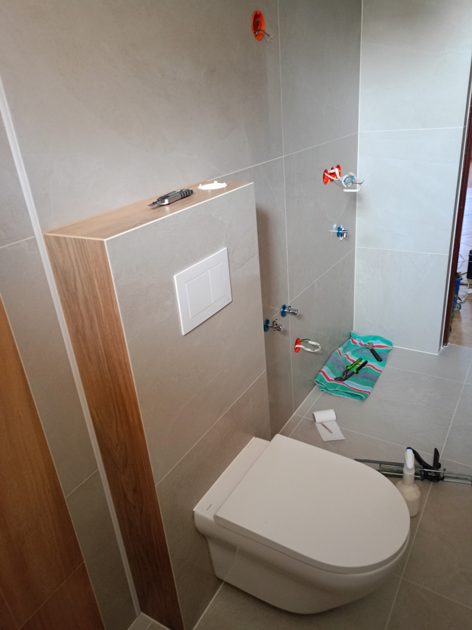 Podwieszana toaleta w trakcie montażu, widoczne narzędzia glazurnicze, szare płytki, instalacja wodna i przyłącza, ręcznik roboczy, częściowo wykończona łazienka w neutralnych kolorach.