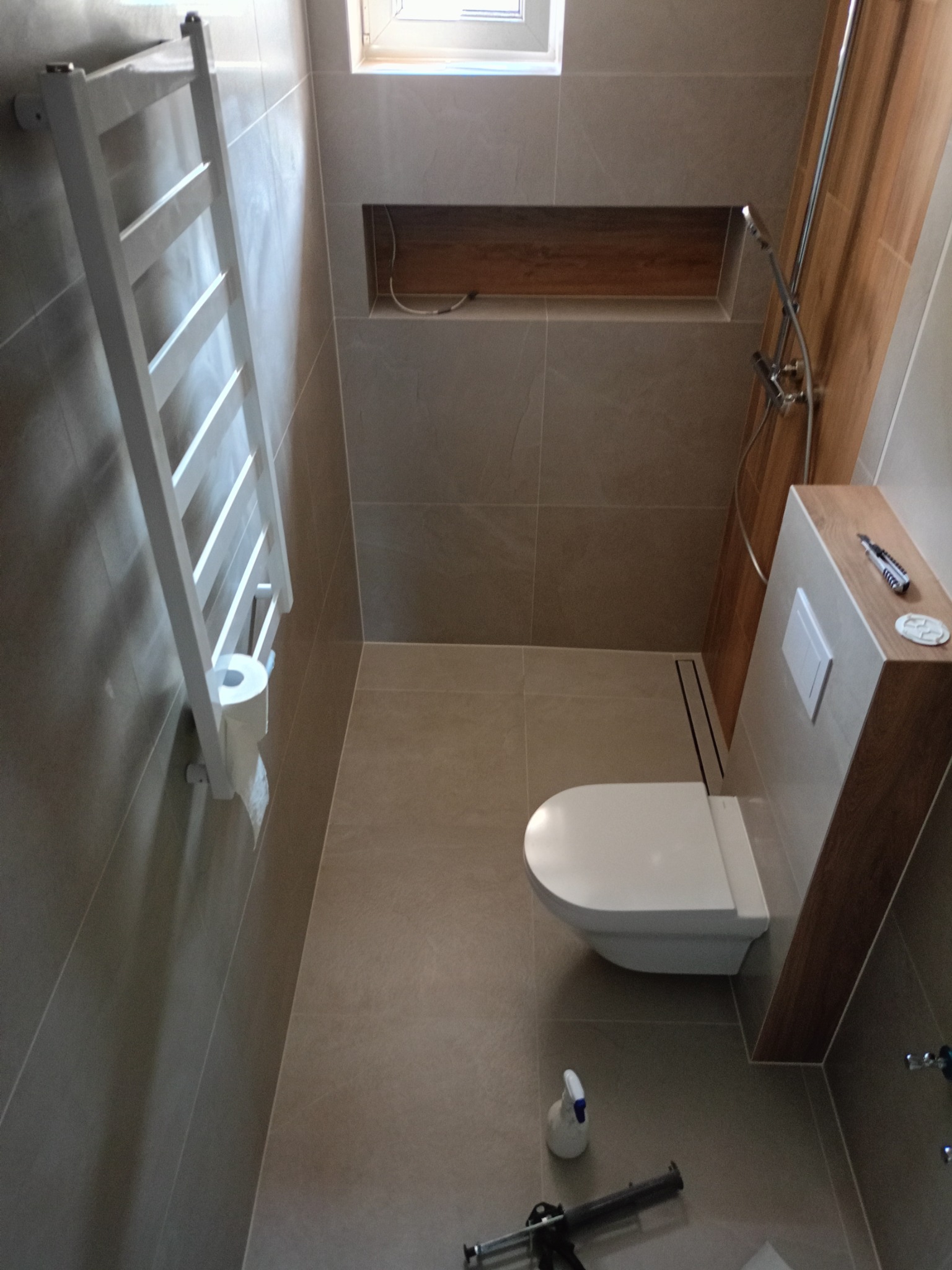 Wykończona łazienka z beżowymi płytkami, wisząca toaleta, grzejnik drabinkowy z papierem toaletowym, wnęka z drewnianym wykończeniem i narzędzia na obudowie spłuczki.