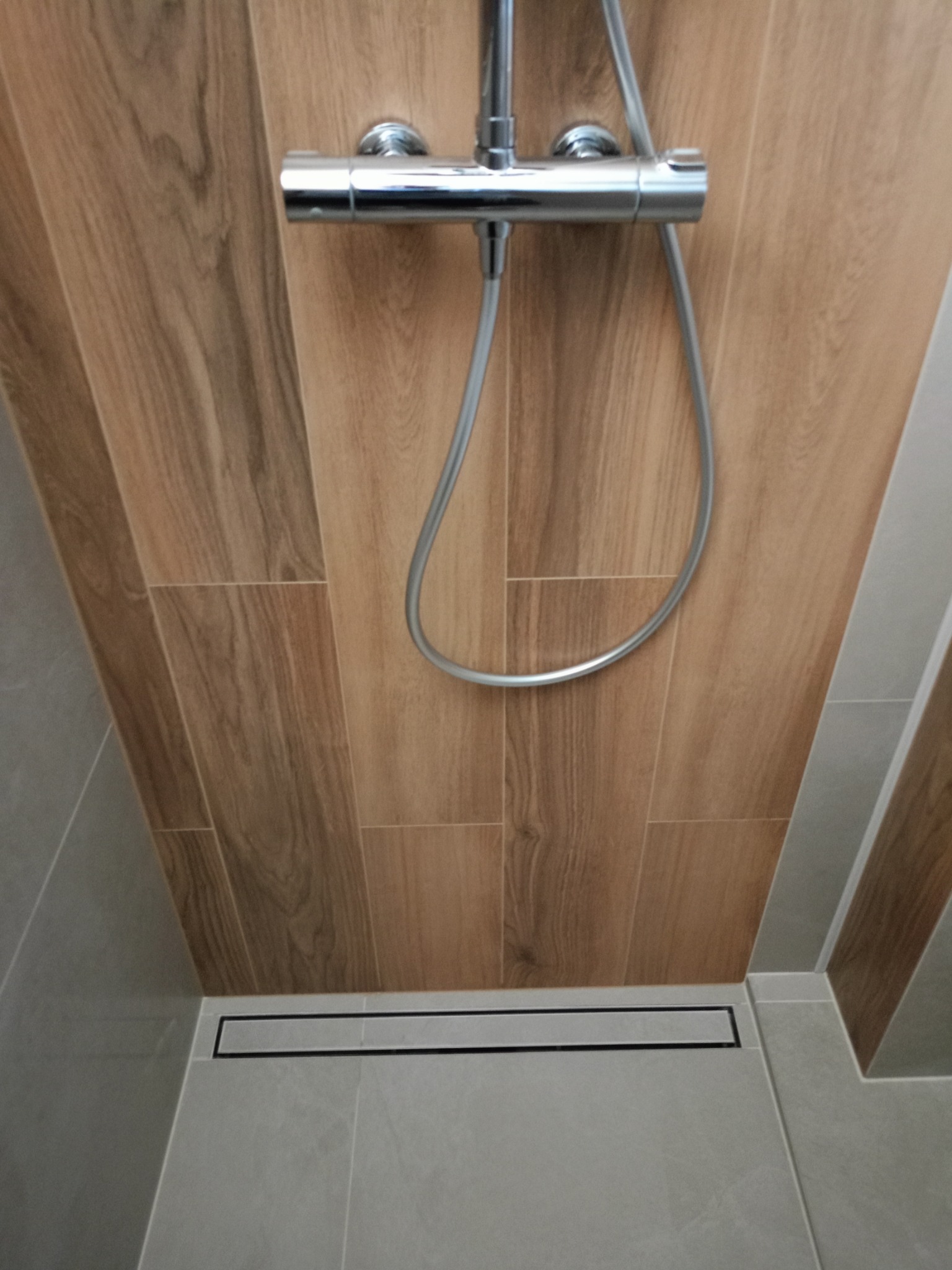 Nowoczesna kabina prysznicowa walk-in z odpływem liniowym i panelami ściennymi imitującymi drewno, chromowana armatura.