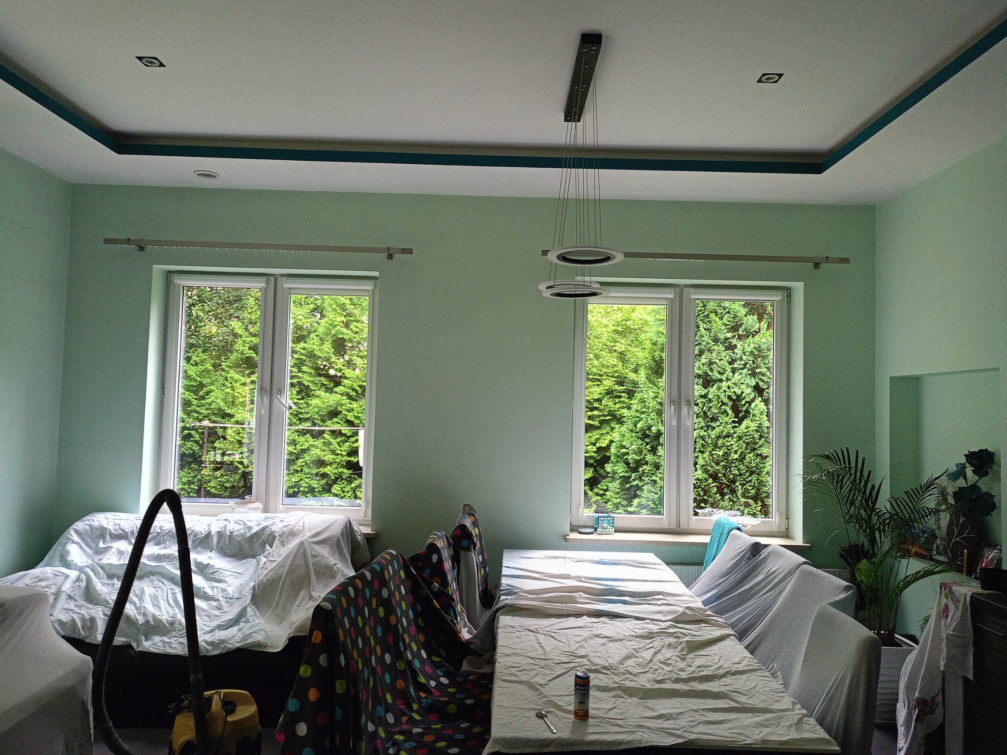 Pokój przed odświeżeniem: ściany w odcieniach zieleni, meble zabezpieczone folią malarską, widoczny odkurzacz i puszka z farbą, okna z widokiem na zieleń.