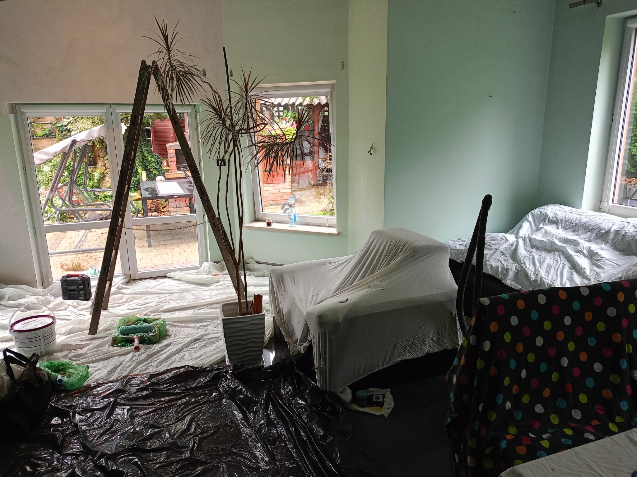 Pokój w trakcie odświeżania: drabina oparta o okno, zabezpieczone meble pod białym materiałem, wiadro z farbą i wałek malarski na folii ochronnej, widoczna pomalowana na zielono ściana.