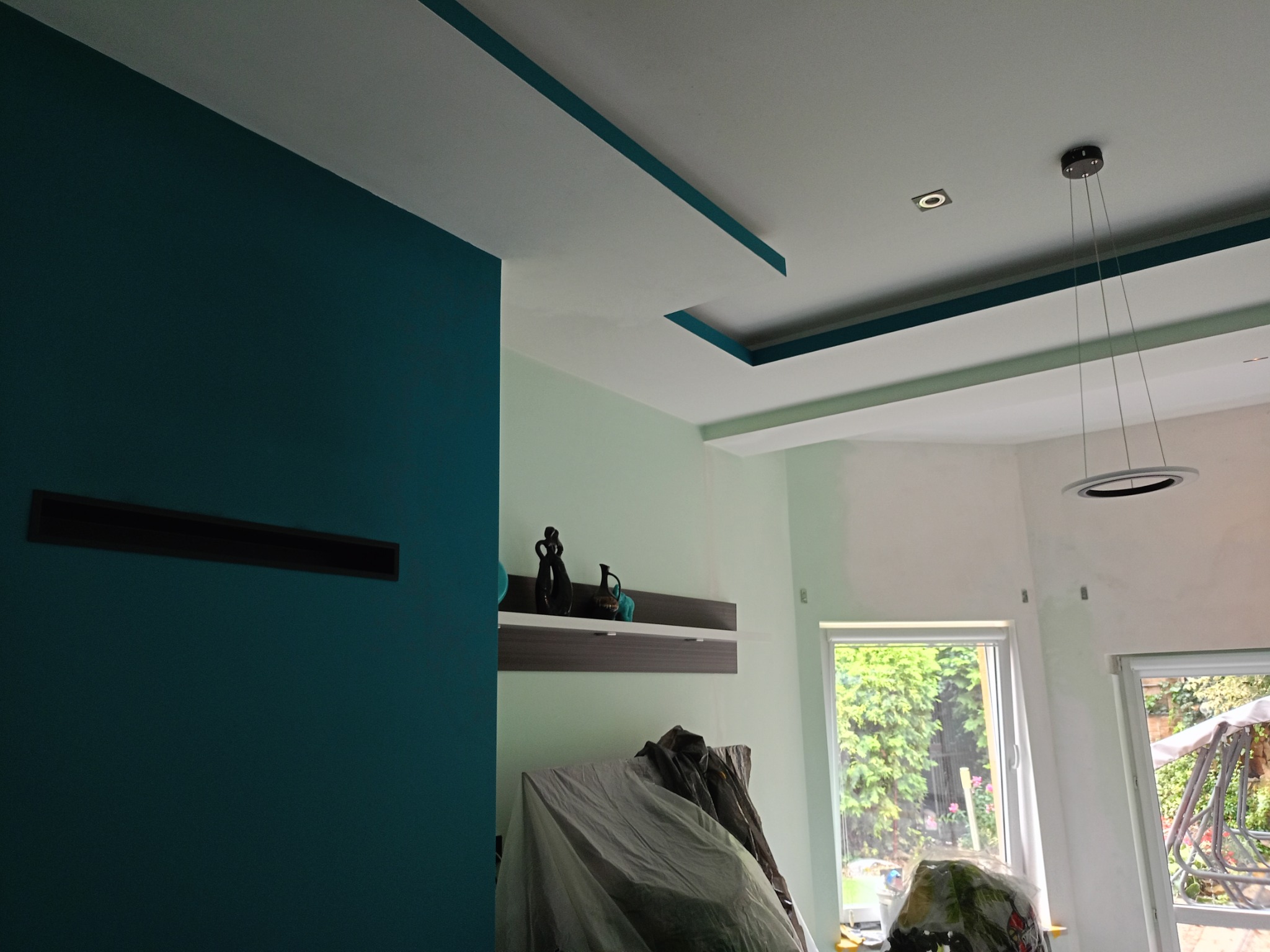 Wnętrze w trakcie prac wykończeniowych: ściana pomalowana na intensywny morski kolor, sufit z dekoracyjnymi wnękami w tym samym odcieniu, zabezpieczone meble folią malarską, widoczna półka...