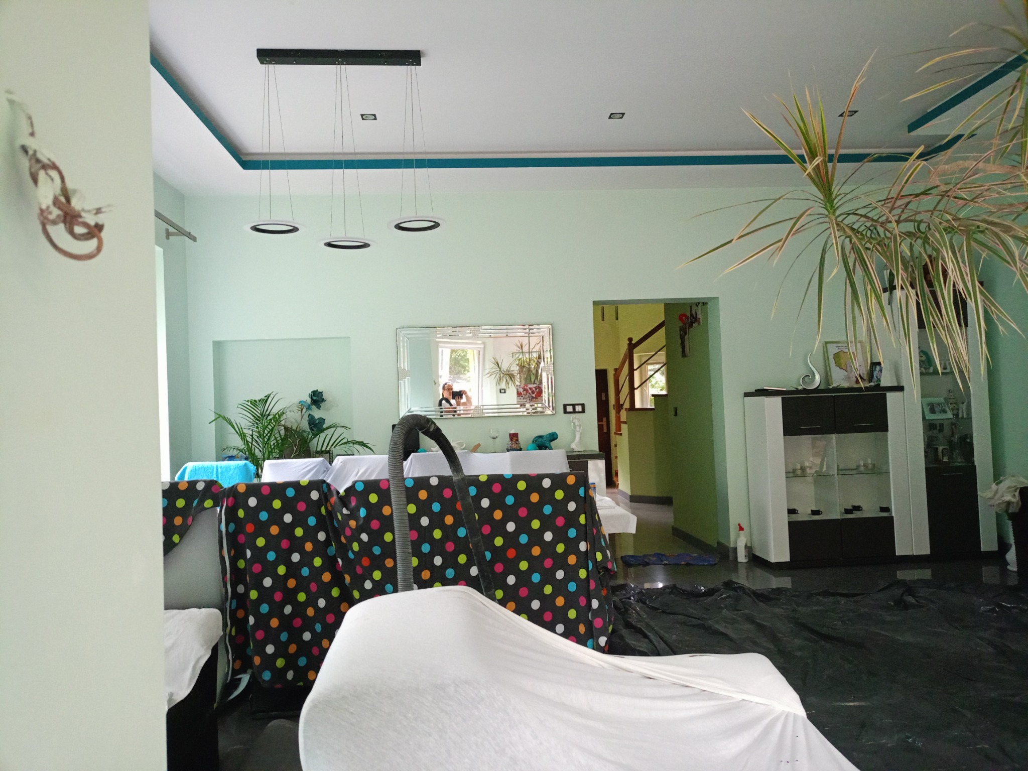 Salon przygotowany do prac malarskich: meble zakryte folią i materiałem ochronnym w kropki, widoczny odkurzacz centralny, podłoga zabezpieczona czarną folią, ściany w kolorze pistacjowym.