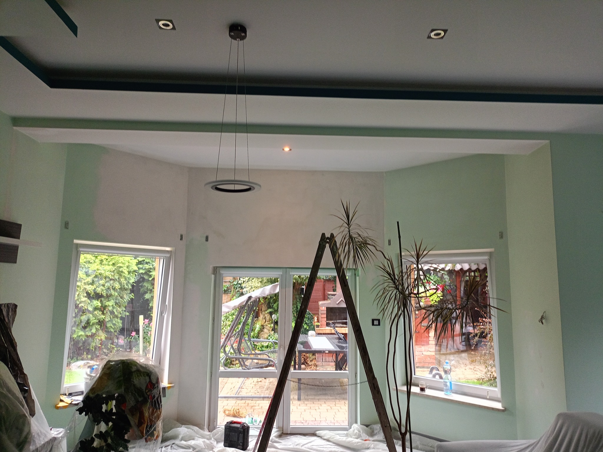 Pokój w trakcie malowania na jasnozielono, zabezpieczony folią malarską, widoczna drabina, okna z widokiem na ogród i nowoczesna lampa wisząca.