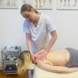 Masaż Legnica Fedorova Studio - Masaż pleców i szyi wykonywany przez masażystkę w białym uniformie, widoczne plakaty anatomiczne na ścianie i wózek z akcesoriami do masażu.