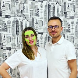 Masaż Legnica Fedorova Studio - Kobieta z zielonymi taśmami kinesiotaping na twarzy, pozująca z mężczyzną na tle tapety w czarno-białe budynki.